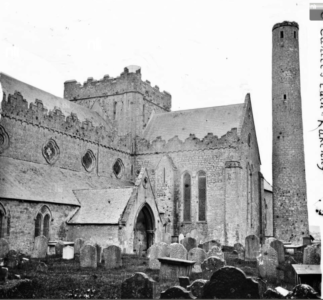 Black and white photo of a stone church with arched windows and the iconic Kilkenny Round Tower. Gravestones fill the foreground, and part of a handwritten vertical label appears on the right side.