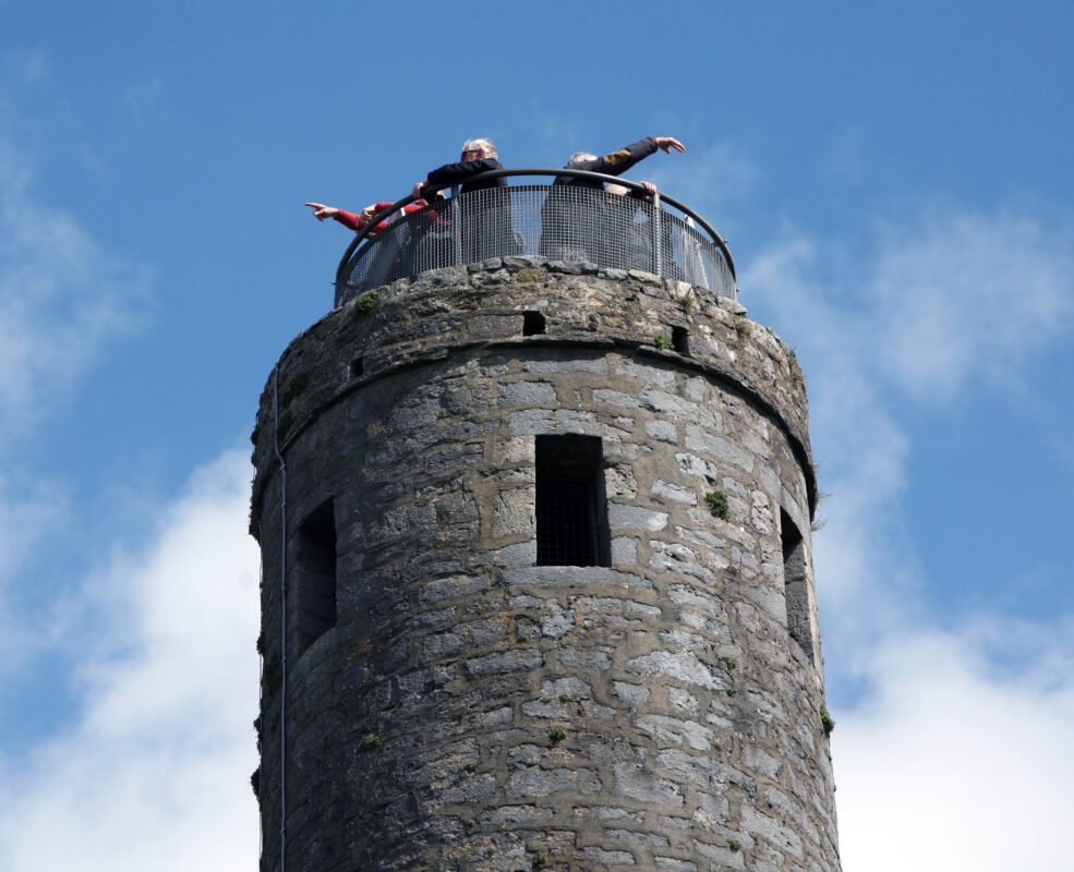 Two people stand on the circular viewing platform of a tall stone tower, leaning over the railing and pointing outward against a background of blue sky and clouds.