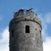 Two people stand on the circular viewing platform of a tall stone tower, leaning over the railing and pointing outward against a background of blue sky and clouds.
