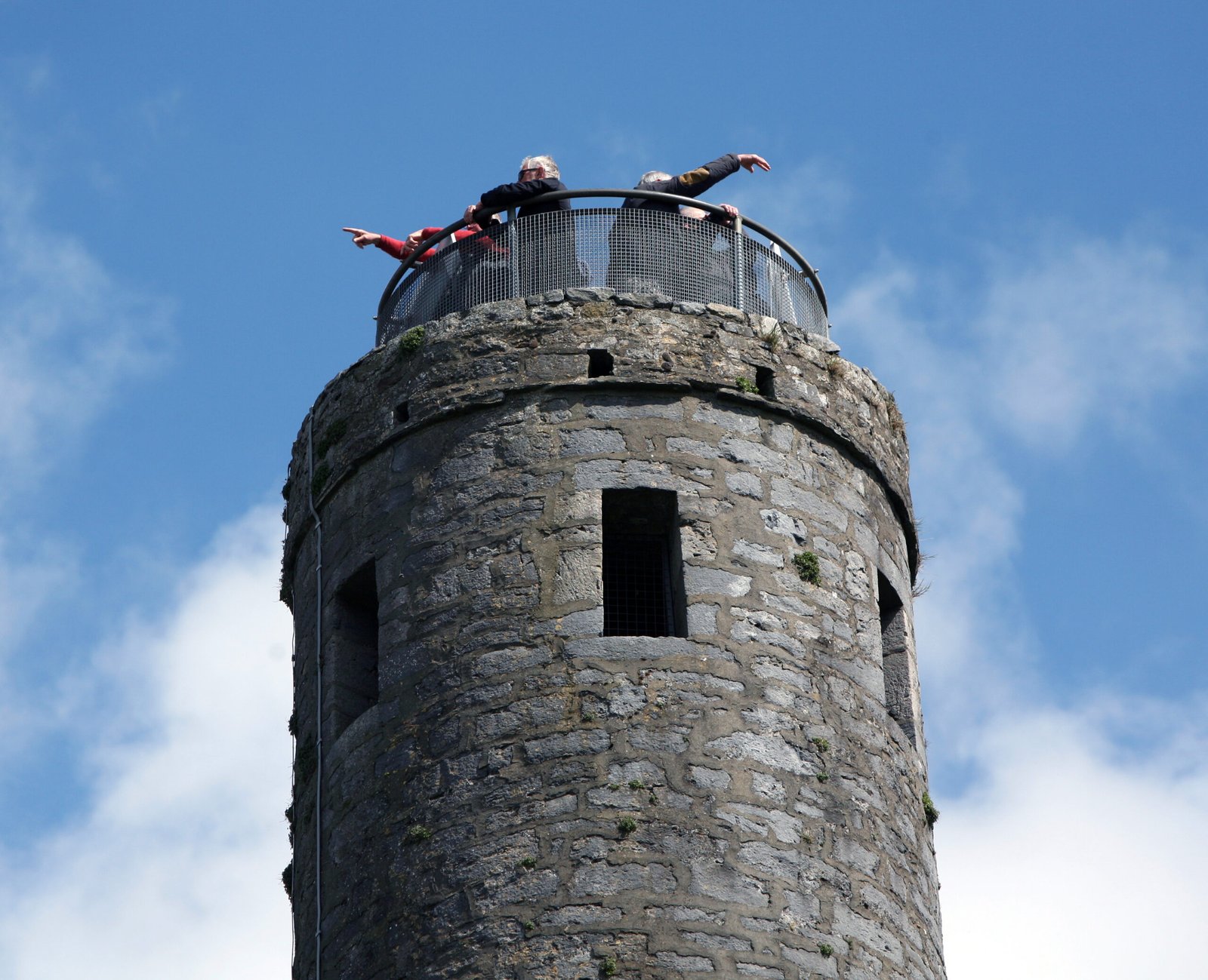 Two people stand on the circular viewing platform of a tall stone tower, leaning over the railing and pointing outward against a background of blue sky and clouds.
