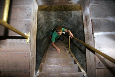 A person in a green shirt climbs up a steep, narrow wooden staircase inside a dimly lit stone tunnel, holding onto a brass handrail for support.