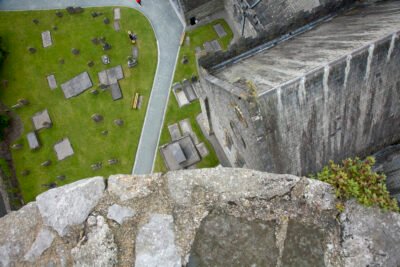 Aerial view from a stone ledge overlooking a church roof and a graveyard with headstones, green grass, pathways, benches, and a few people walking below.