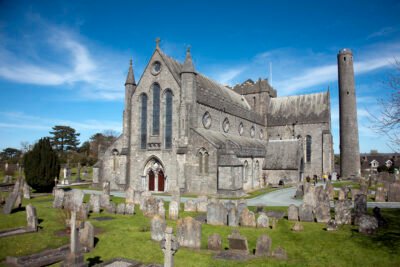 A historic stone cathedral with pointed arches and stained glass windows stands among old gravestones and monuments in a grassy cemetery, beneath a clear blue sky. A tall round tower rises beside the church.