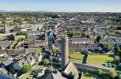 Aerial view of a town with a prominent round tower in the center, surrounded by historic buildings, houses, greenery, and distant hills under a clear sky.